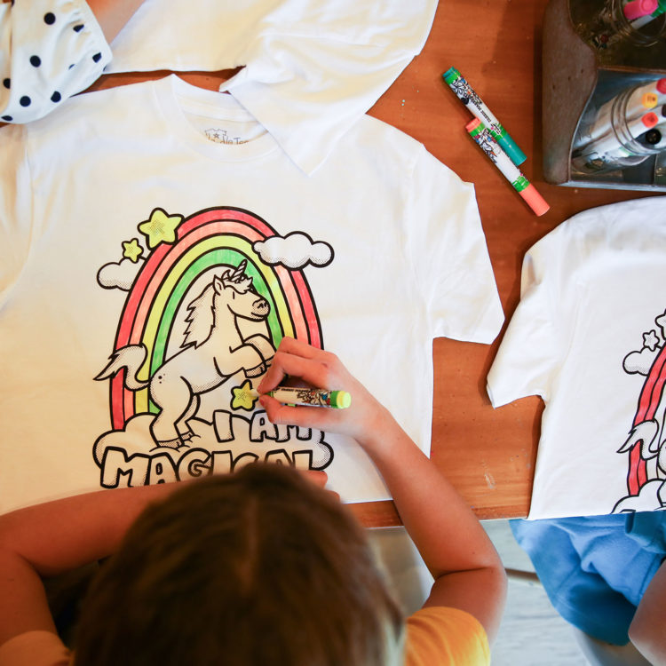 "Photo of kid using fabric markers to color the Doodle Tees 'I Am Magical" design printed on a shirt"