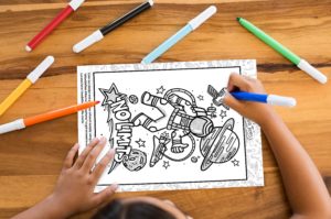 "Photo of a kid coloring with markers Doodle Tees' 'No Limits' astronaut design printed on a paper"